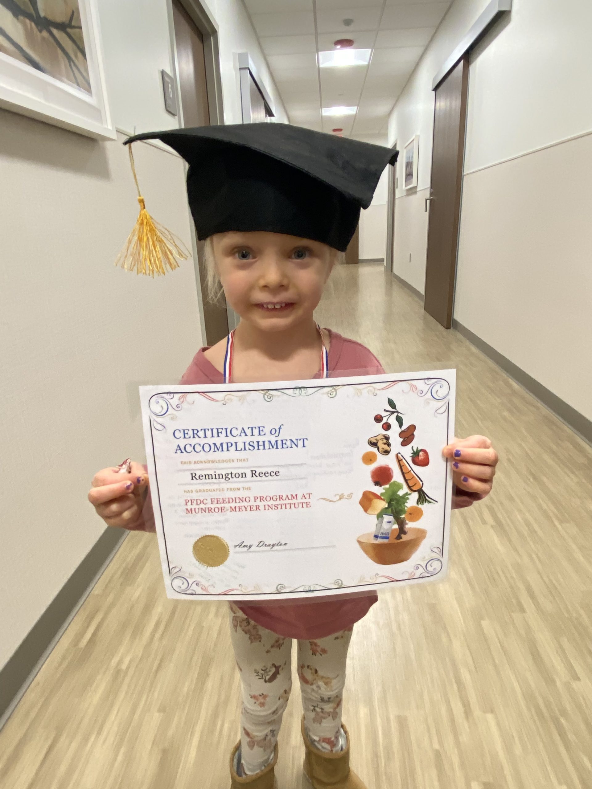 Remi stands holding her graduation certificate while wearing a graduation cap and a big smile